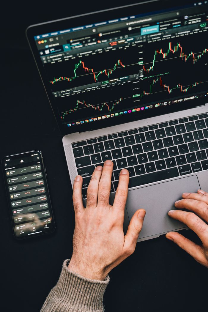 gallery-1 Hands typing on a laptop with cryptocurrency charts and smartphone display in view, focusing on digital finance.